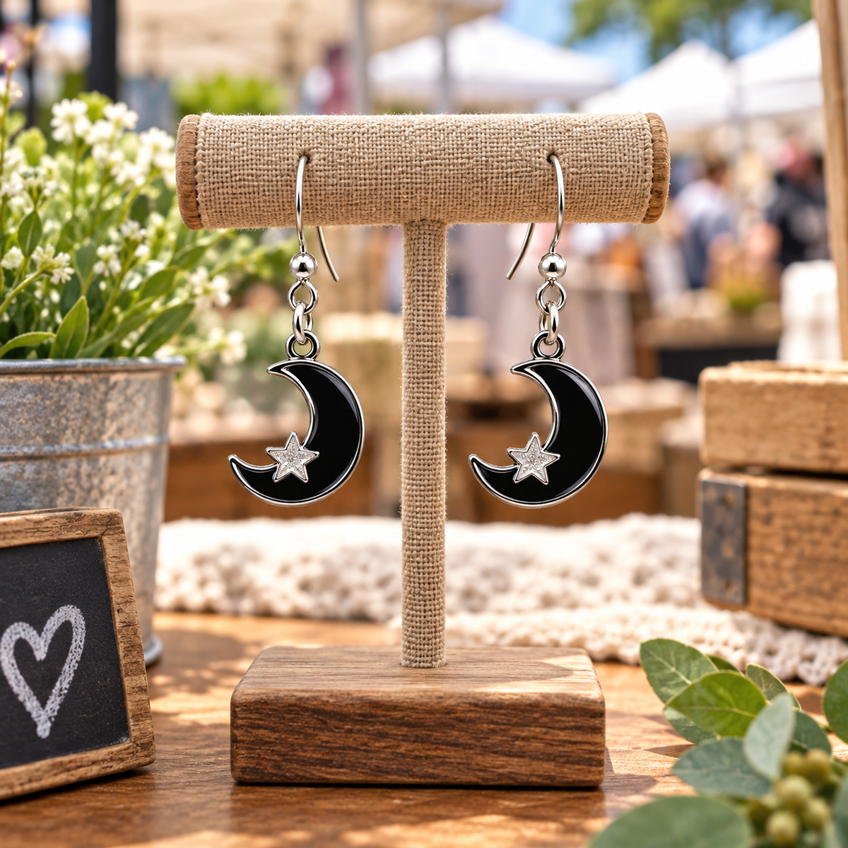A pair of black crescent moon and star drop earrings with silver hooks, displayed on a burlap-wrapped jewelry stand on a wooden table in an outdoor market setting.