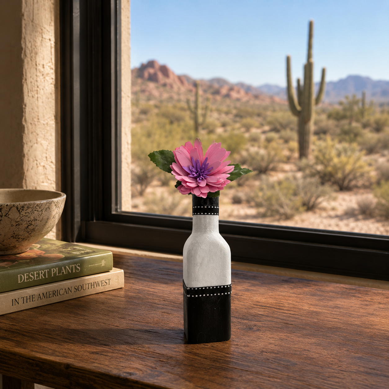 Vase with a pink flower on a wooden table in front of a window with a desert view.