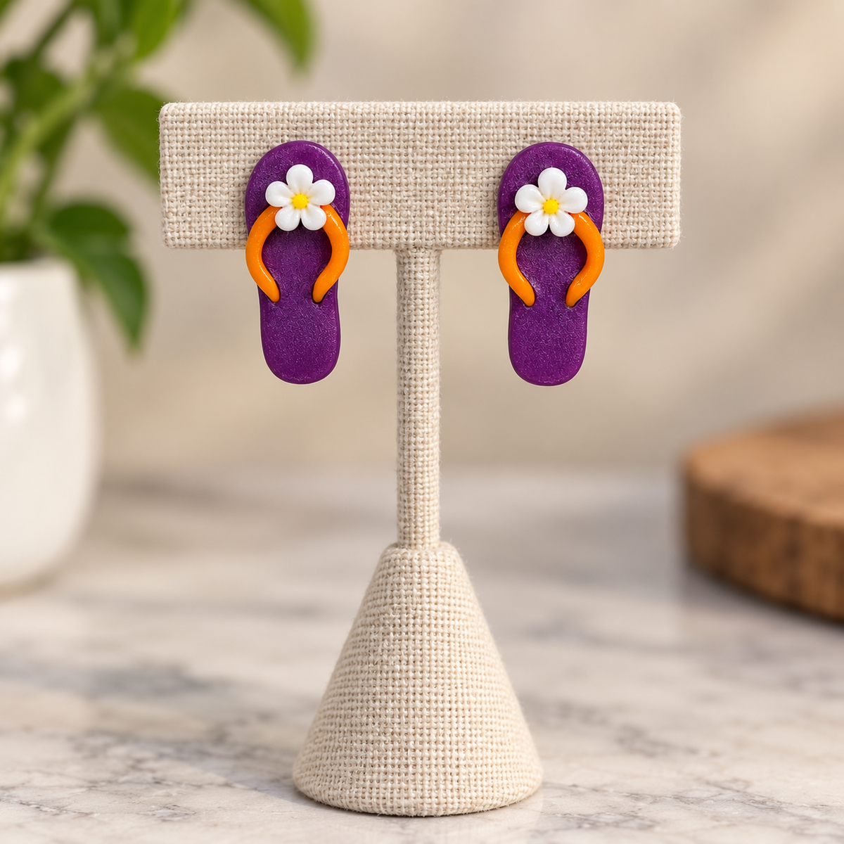 A pair of purple flip-flop earrings with orange straps and white daisy flower charms, displayed on a beige woven jewelry stand.