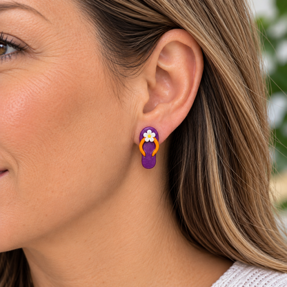 A close-up of a woman's ear wearing a small flip-flop-shaped stud earring in purple and orange with a white flower detail, against a softly blurred background.