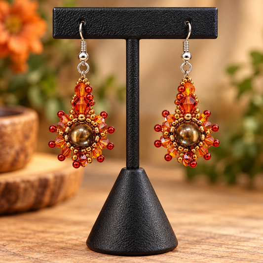 A pair of handmade beaded dangle earrings displayed on a black jewelry stand. The earrings feature a central round brown bead surrounded by radiating red and orange beads with small metallic gold-toned beads, hanging from silver fishhook wires.