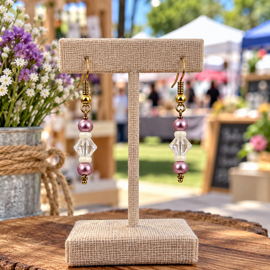 A pair of handmade dangle earrings displayed on a burlap-covered jewelry stand at an outdoor market. The earrings feature gold-tone metal hooks, white beads, faceted clear crystal beads, and pink pearl-like beads.