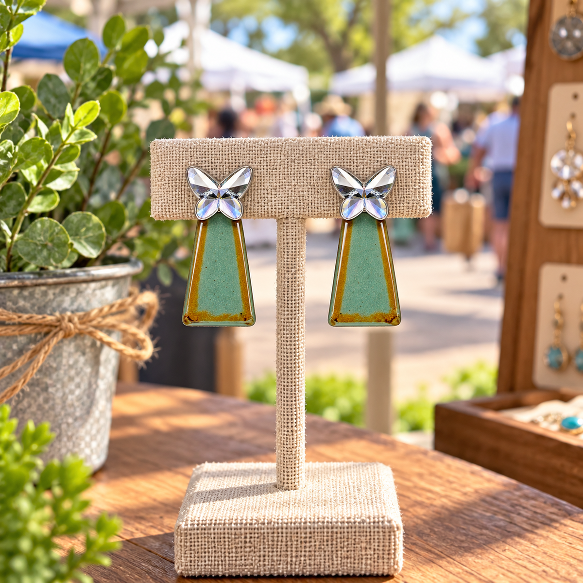 A faceted silver-colored butterfly with a teal-green geometric drop accented with warm golden tones on a display stand