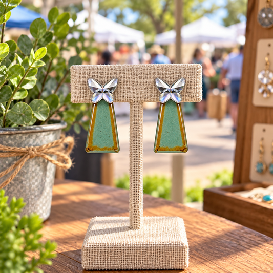 A faceted silver-colored butterfly with a teal-green geometric drop accented with warm golden tones on a display stand