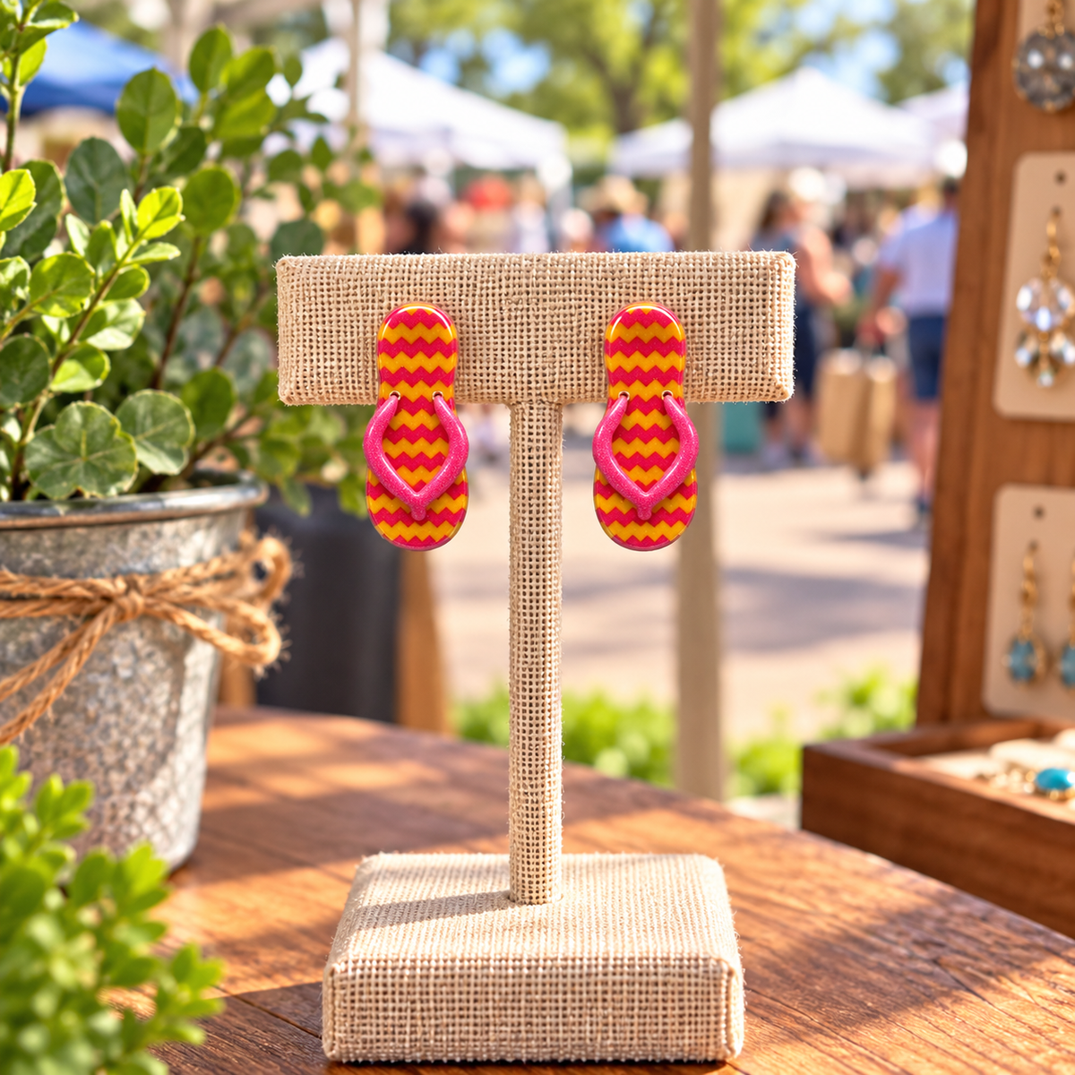 A pair of flip-flop style earrings with pink and yellow chevron-patterned tops and pink straps, displayed on a burlap-covered jewelry stand at an outdoor market.